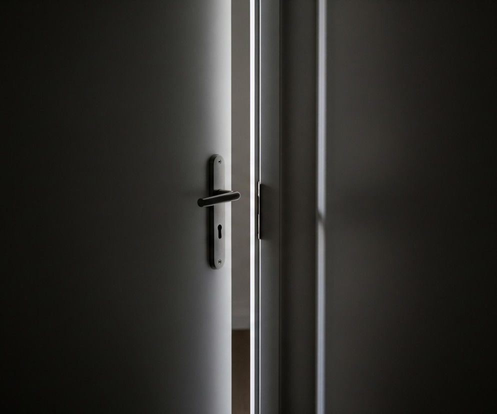 Półotwarte, ciemne drzwi zajmują centrum kadru, z wyraźnym metalowym uchwytem klamki i otworem na klucz na ich powierzchni. Wąska, pionowa szczelina światła rozciąga się od góry do dołu między drzwiami a ciemną futryną po prawej stronie. Otoczenie drzwi po lewej i prawej stronie jest pogrążone w głębokim cieniu, co kontrastuje z jasnym, rozproszonym światłem wpadającym przez szczelinę.