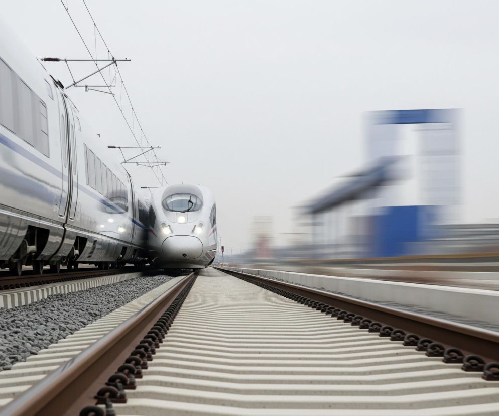 Dwa nowoczesne pociągi dużych prędkości, jeden jadący z lewej strony, drugi z prawej, na szarym tle, symbolizujące rozwój transportu i inwestycje w infrastrukturę kolejową związane z Portem Polska, o czym czytać można na Super Biznes.