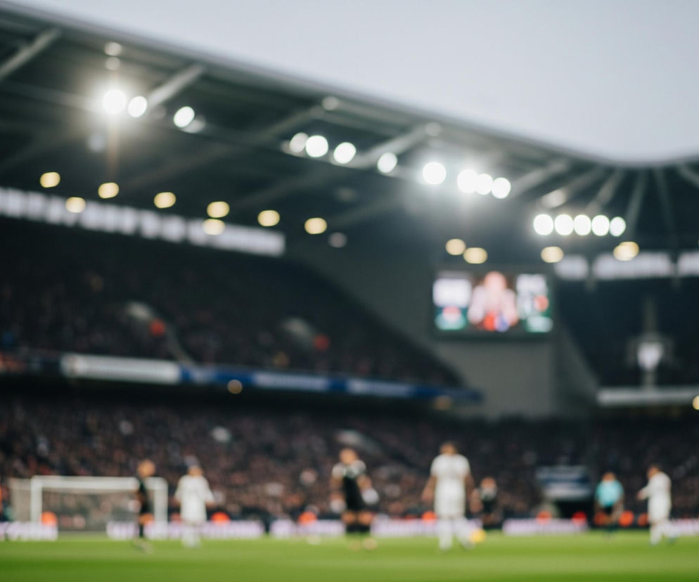 Rozmazany obraz przedstawia stadion piłkarski oświetlony jasnymi reflektorami. Na dole widoczne jest zielone boisko, a na nim niewyraźne sylwetki graczy w strojach sportowych, głównie białych, czarnych i pomarańczowych. W tle rozpościera się ciemna trybuna wypełniona niezidentyfikowanym tłumem widzów, nad którą widać jasne lampy oświetlające całą przestrzeń. Po prawej stronie w głębi znajduje się duży ekran, na którym wyświetlane są niewyraźne obrazy.