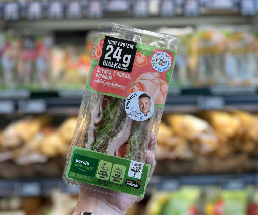 Dłoń osoby trzymającej kanapkę Tomcio Paluch na tle sklepowych regałów. O ofercie sieci Żabka przeczytasz na Eska.