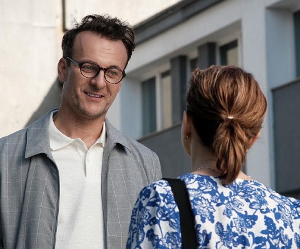 Radomir (Łukasz Wójcik) w okularach i koszuli polo rozmawia z Dorotą Kujawiak (Karina Seweryn), stojącą tyłem, w kwiecistej bluzce. To scena z serialu Barwy Szczęścia, gdzie Radomir planuje okłamać Dorotę, o czym przeczytasz na Super Seriale.