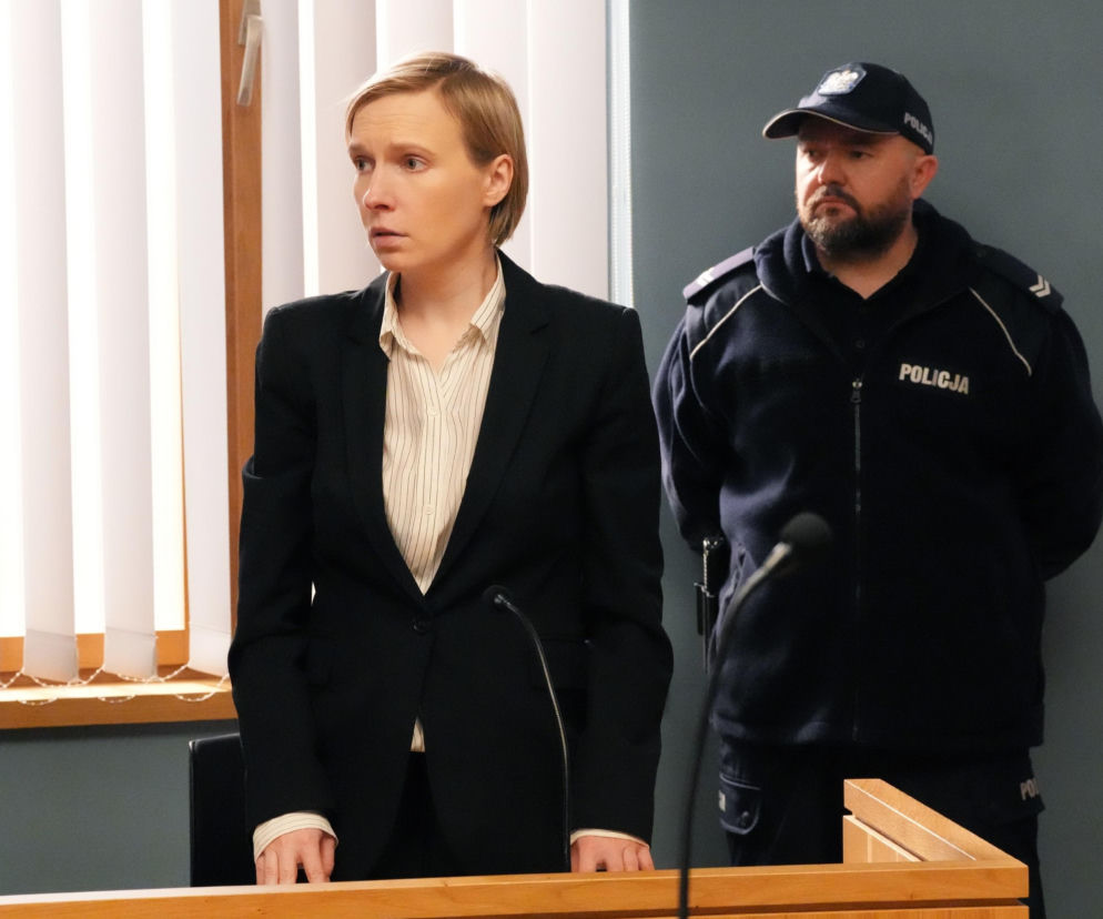 Zmartwiona Natalia Zwoleńska (Maria Dejmek) stoi w czarnym garniturze z paskowaną koszulą za drewnianym pulpitem w sali sądowej, obok niej policjant z zarostem i w czapce z napisem POLICJA. Scena z serialu Barwy szczęścia dostępna na Super Seriale.