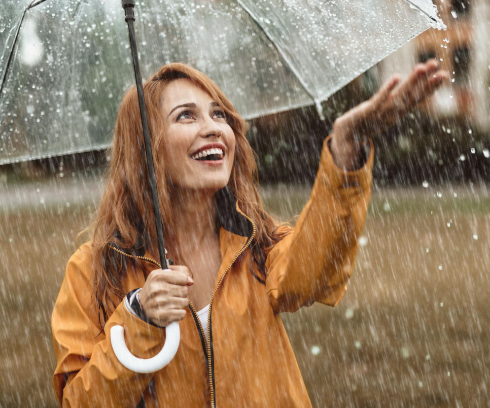 Młoda kobieta o rudych włosach w żółtej kurtce radośnie uśmiecha się, trzymając parasol i wystawiając rękę, by poczuć padający deszcz. Deszczowe krople wirują wokół niej, tworząc przyjemną, relaksującą atmosferę, o której można przeczytać na portalu Poradnik Zdrowie.