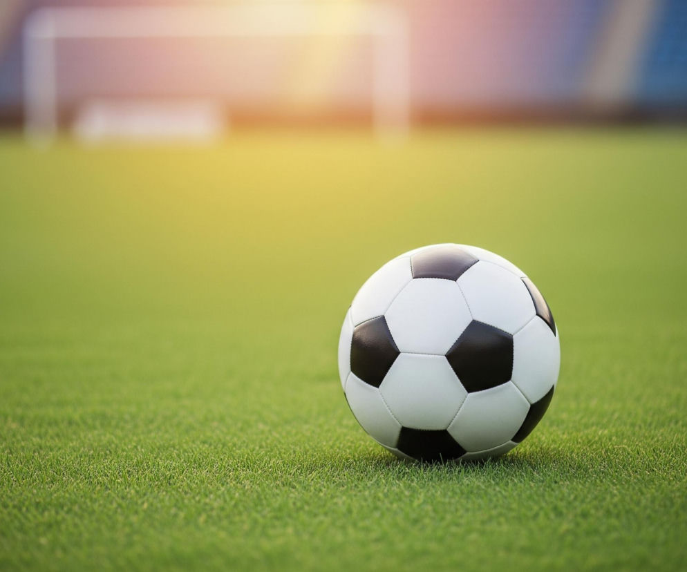 Czarno-biała piłka nożna leży na jasnozielonym, krótkim trawiastym boisku. Piłka jest umieszczona na środku kadru, bliżej dołu, a jej powierzchnia jest wyraźnie widoczna, ukazując heksagonalne i pentagonalne panele. W tle, rozmyte obiekty to bramka piłkarska z białą konstrukcją, oraz części trybun w odcieniach niebieskiego. Nad bramką i częścią boiska widać jasne, rozmyte światło, przypominające zachodzące słońce lub sztuczne oświetlenie.