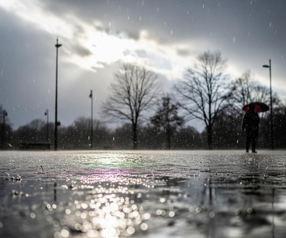 Na pierwszym planie, w dolnej części obrazu, znajduje się mokra, błyszcząca powierzchnia, prawdopodobnie asfaltowa lub betonowa, z widocznymi odbiciami światła, tworzącymi efekt bokeh. Powierzchnia ta jest pokryta licznymi kroplami deszczu, które rozbryzgują się delikatnie, tworząc drobne wzniesienia. W tle, pod pochmurnym, szarym niebem z prześwitami jaśniejszego słońca, widać trzy niewyraźne, ciemne sylwetki ludzi, z których dwie trzymają parasole – jeden czarny, a drugi częściowo czerwony. Sylwetki drzew bez liści i wysokie słupy oświetleniowe są rozmieszczone w tle, dodając kontekst miejski lub parkowy.