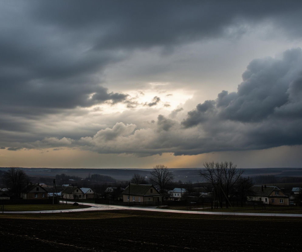 Na obrazie dominują ciemne, burzowe chmury, które pokrywają większą część nieba, z prześwitem jaśniejszego, lekko żółtawego światła słonecznego w centralnej części. Poniżej rozciąga się wiejski krajobraz z widocznymi budynkami mieszkalnymi o jasnych elewacjach i ciemnych dachach, rozmieszczonymi wzdłuż wijącej się drogi. Przed domami widoczne są puste pola uprawne w kolorze ciemnobrązowym, a na horyzoncie zarysowuje się zalesiony obszar i pagórkowaty teren, wszystko spowite w półmroku i chłodnych barwach.