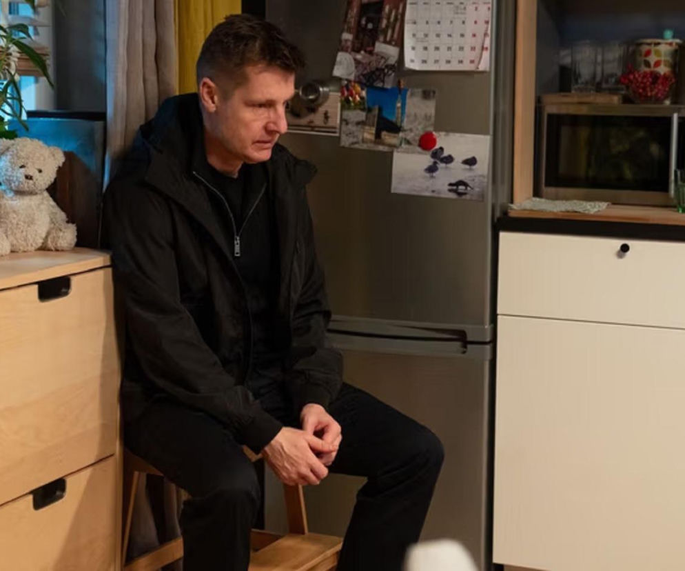 Paweł Zduński (Rafał Mroczek) w czarnej kurtce i spodniach, siedzi smutny na krześle, patrząc w lewo. Tło przedstawia wnętrze kuchni z szafkami i lodówką z przypiętymi zdjęciami. Ta scena z M jak miłość to zapowiedź dramatycznych wydarzeń. Więcej o losach bohatera przeczytasz na portalu Super Seriale.
