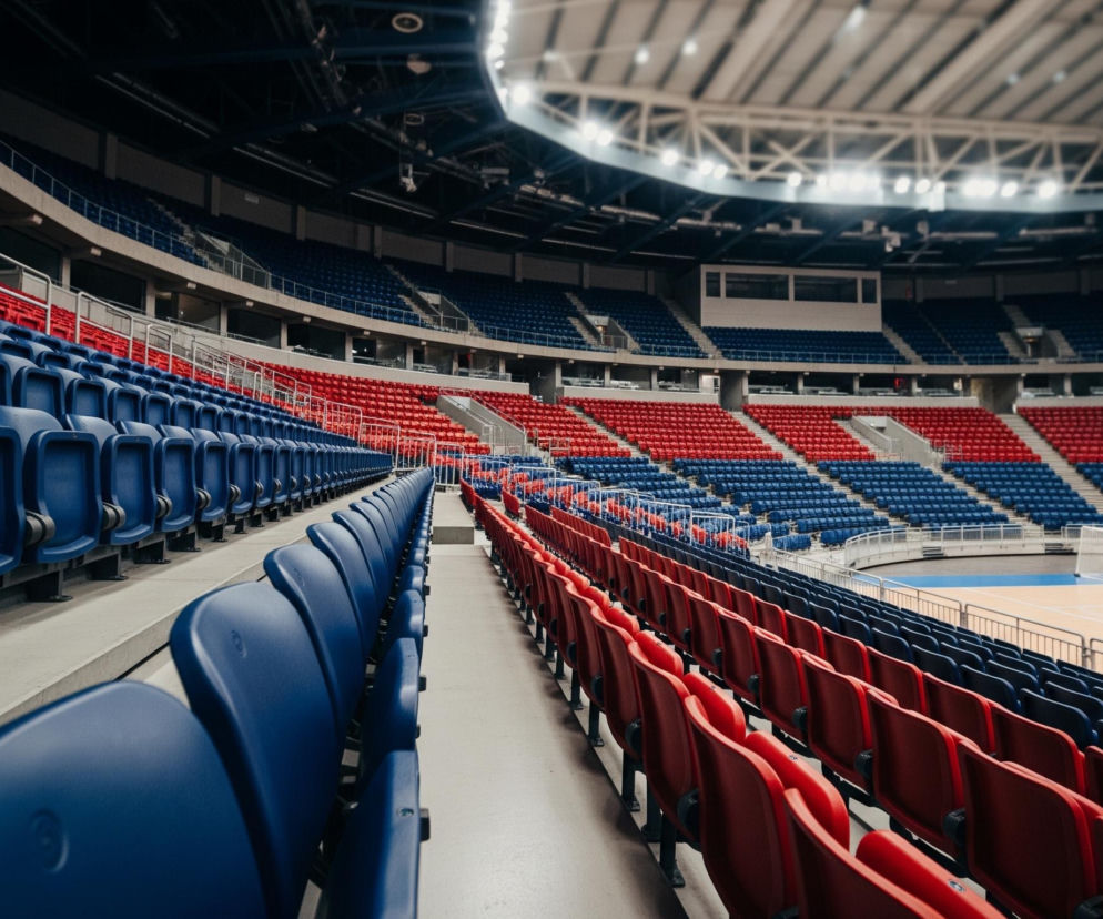 Wnętrze pustej hali sportowej oświetlone jasnymi światłami pod sufitem, przedstawione z perspektywy rzędu siedzeń. Obraz skupia się na długich rzędach pustych, plastikowych krzesełek w kolorach ciemnoniebieskim i czerwonym, biegnących wzdłuż trybun. Po lewej stronie dominuje rząd niebieskich siedzeń, natomiast po prawej widoczne są czerwone siedzenia, tworzące wyraźny kontrast. W tle znajduje się boisko z drewnianym parkietem z jasnoniebieskimi pasami, a także bramka z biało-czerwonymi słupkami i białą siatką.