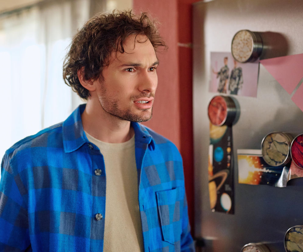 Mężczyzna z kręconymi włosami w niebieskiej koszuli w kratę i beżowym podkoszulku, patrzący w bok z zaniepokojeniem, stoi przy lodówce ozdobionej magnesami i notatkami. Scena z serialu Na Wspólnej, pełna napięcia. Więcej informacji o serialu znajdziesz na Super Seriale.