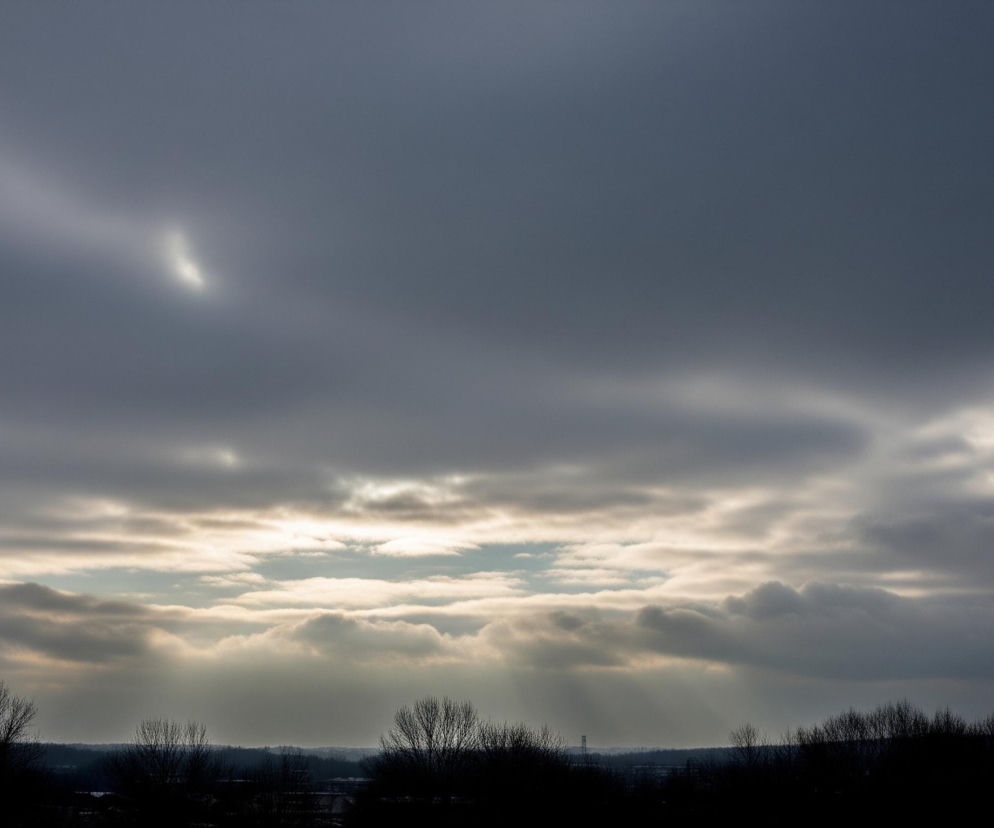Ciemne, szare chmury dominują niebo, tworząc dynamiczny kontrast z jaśniejszymi fragmentami, przez które przebija się błękitne niebo w centrum kadru. Od dołu ciemne sylwetki bezlistnych drzew i krzewów tworzą wyraźną granicę z horyzontem. Między chmurami widoczne są promienie słoneczne, które rozświetlają centralną część obrazu, tworząc efekt poświaty.