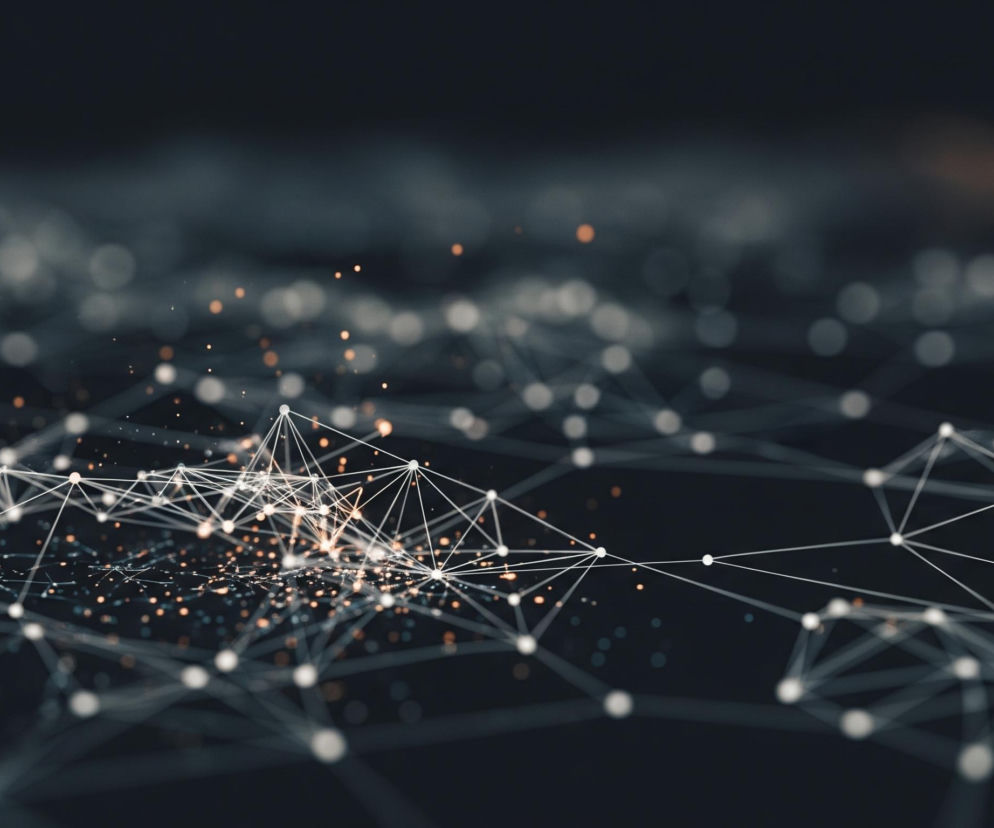 Gęsta sieć białych linii łączy liczne jasne punkty, tworząc trójwymiarową strukturę w centrum obrazu. Centralny obszar sieci rozświetla intensywny blask oraz liczne złociste iskierki, rozpraszające się wokół punktów połączeń. Ciemnoszare tło wypełniają rozmyte, owalne światła i delikatne linie, układające się w podobną, lecz mniej wyraźną siatkę, zanikającą w oddali.