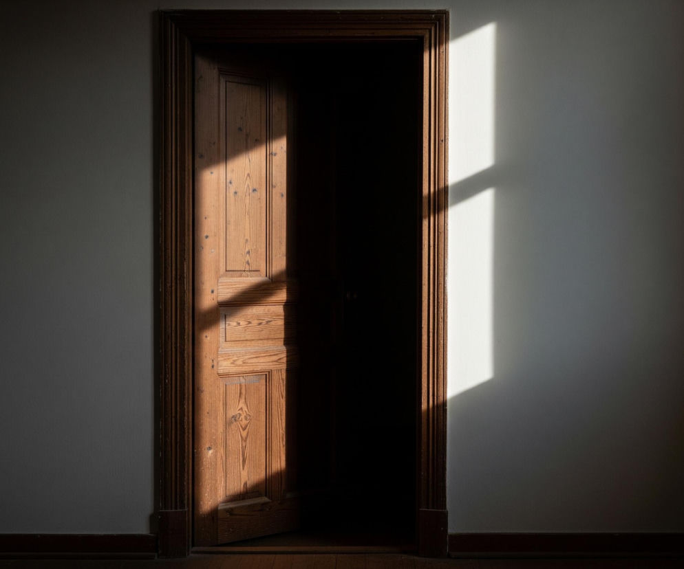 Drewniane, uchylone drzwi z wyraźnym usłojeniem zajmują centralną część kadru, częściowo oświetlone ukośnym pasmem światła słonecznego padającym z lewej strony. Lewa połowa drzwi oraz fragment ościeżnicy są jasne, podczas gdy prawa strona i wnętrze otworu drzwiowego pozostają w głębokim cieniu. Jasnoszare ściany otaczają ościeżnicę, z prawą ścianą również częściowo oświetloną pionowym prostokątem światła, rzucającym cień na kształt krzyża w górnej części. Na dole widoczna jest ciemnobrązowa listwa przypodłogowa i fragment drewnianej podłogi.