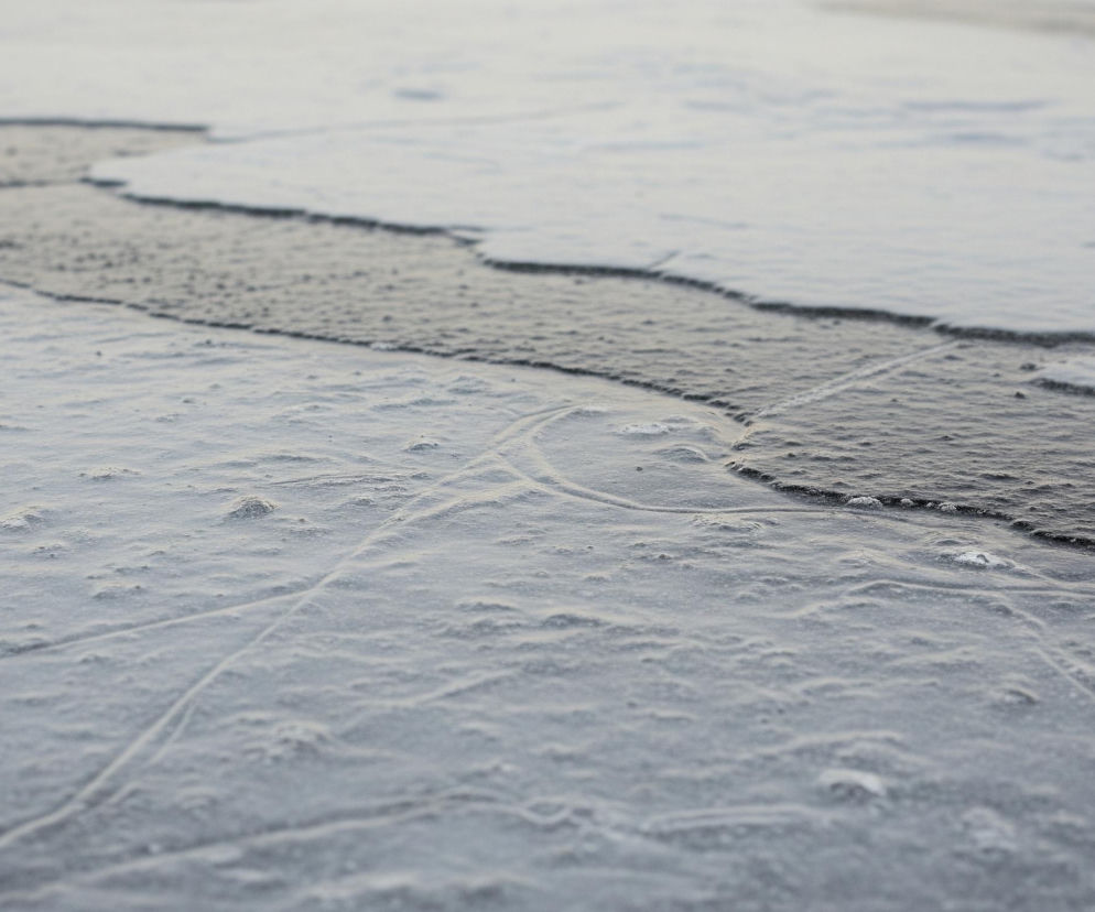 Zmarznięta powierzchnia zajmuje większą część kadru, z wyraźną linią brzegową oddzielającą ją od ciemniejszej, płynnej wody na górze. Lód jest nieregularny, z małymi wypukłościami i subtelnymi, zakrzywionymi śladami na przedzie. Woda przy brzegu wydaje się lekko zmarszczona, tworząc cienkie, białe fale.
