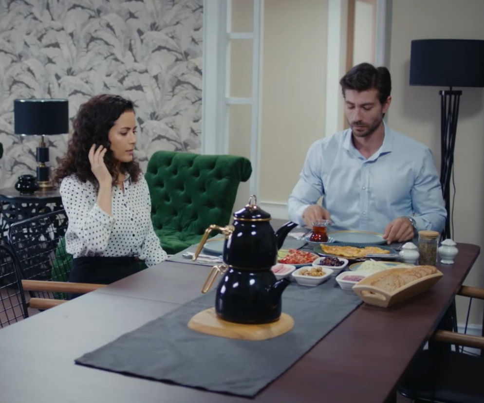 Kobieta z kręconymi włosami i mężczyzna w błękitnej koszuli siedzą przy stole, jedząc śniadanie. Pomiędzy nimi znajduje się czarny dzbanek do herbaty, a na stole widoczne są różne przystawki i pieczywo. Scena oddaje atmosferę wspólnego posiłku z serialu Dziedzictwo, o którym więcej przeczytasz na Super Seriale.