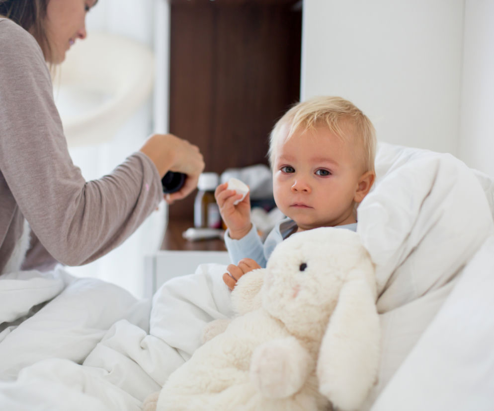 Chory chłopiec w łóżku z maskotką. Obok mama przygotowuje leki. O objawach trzeciego migdałka przeczytasz na SE.