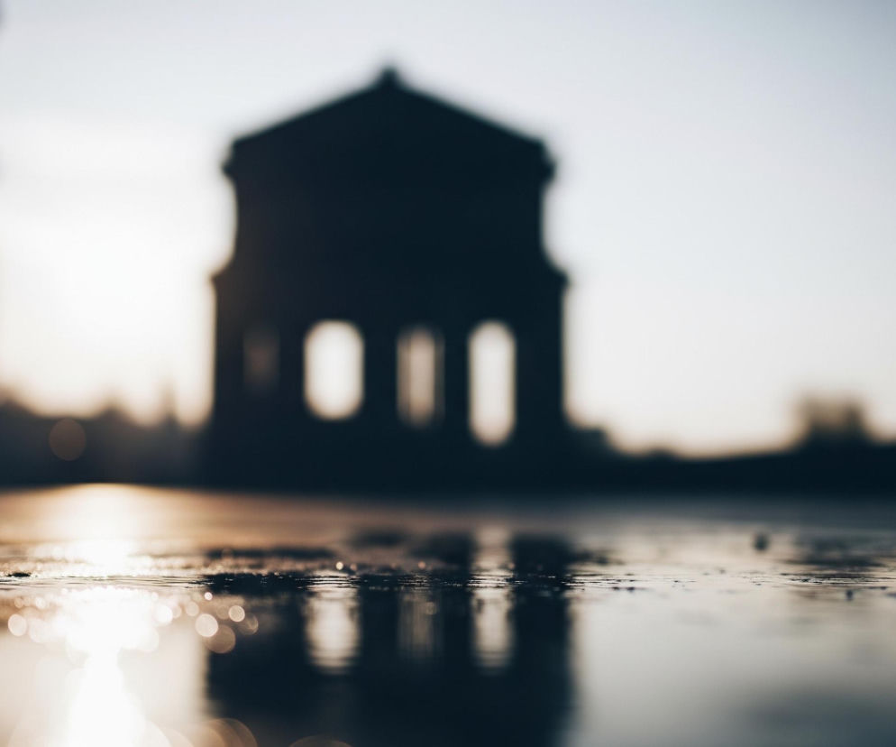 Na pierwszym planie, w dolnej części kadru, znajduje się mokra, odbijająca światło powierzchnia, prawdopodobnie asfaltowa nawierzchnia ulicy lub placu. Widoczne są liczne, drobne krople wody i odbicia jasnego światła, tworzące efekt bokeh. W oddali, w centrum kadru, widać rozmyty zarys ciemnego budynku o klasycznej architekturze, z widocznymi kolumnami lub łukami w dolnej części, a także inne, niewyraźne, ciemne sylwetki budynków po lewej stronie. Tło to jasne, rozmyte niebo, które przechodzi od bieli i jasnego szarości w dolnej części do ciemniejszego błękitu w prawym górnym rogu.