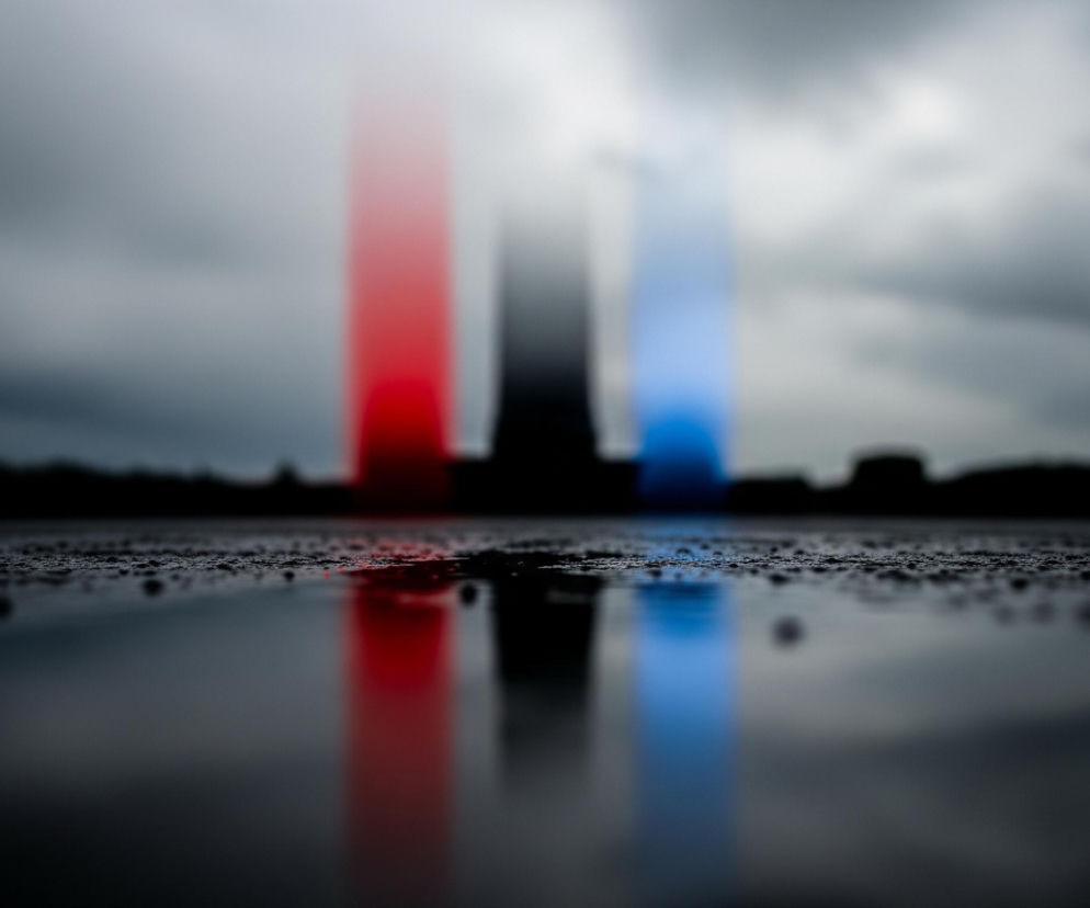 Na pierwszym planie widoczna jest mokra powierzchnia, pokryta licznymi drobnymi kropelkami wody, która odbija światło i barwne smugi. Po lewej stronie odbija się czerwona pionowa smuga, a po prawej niebieska, obie rozmyte i wydłużone, wskazujące na źródła światła poza kadrem lub na horyzoncie. Tło to ciemne, zachmurzone niebo z lekko jaśniejszym obszarem w centrum, oraz sylwetki niewyraźnych, ciemnych budynków lub drzew na horyzoncie.
