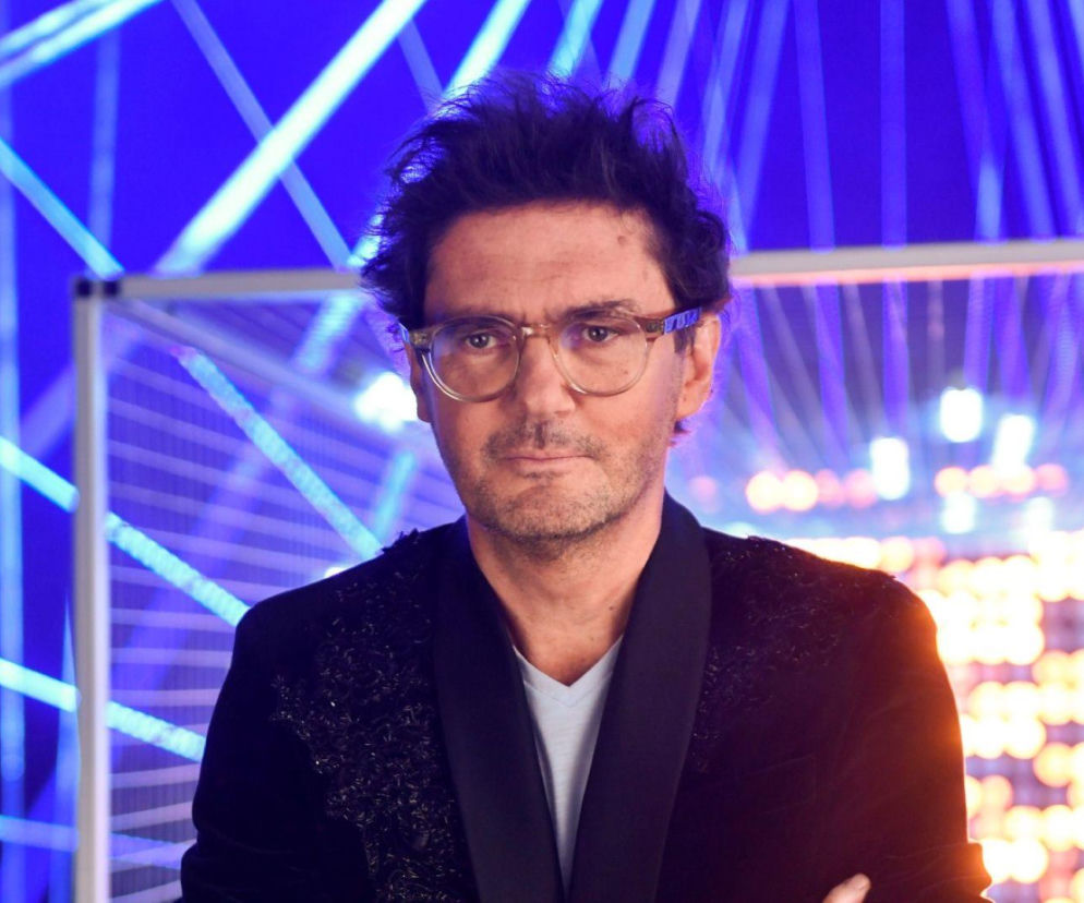 Kuba Wojewódzki w ciemnej marynarce i okularach na tle neonowych świateł. O jego metamorfozie przeczytasz na SE.