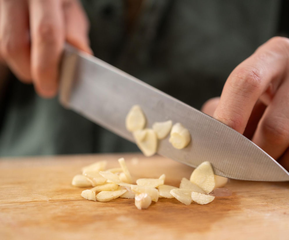 Ręce krojące ząbki czosnku na drewnianej desce, ostry nóż w dłoniach, a obok świeżo posiekane kawałki. Czosnek ceniony za wpływ na zdrowie serca i profilaktykę zakrzepicy, o czym przeczytasz więcej na Poradnik Zdrowie.