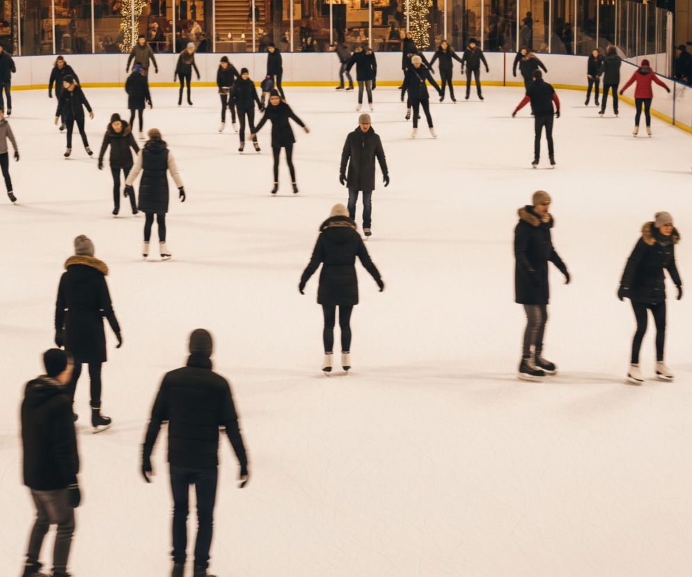 Duża grupa ludzi na lodowisku ślizga się na łyżwach. Większość osób ubrana jest w ciemne, zimowe kurtki i spodnie, z kilkoma wyjątkami w jasnych lub kolorowych okryciach wierzchnich, takich jak beżowa kurtka czy niebieska pikowana kurtka. Lodowisko, otoczone niską bandą w górnej części kadru, ma białą, gładką powierzchnię. W tle, za bandą, widoczne są budynki z oświetlonymi oknami i dekoracjami świątecznymi, tworzącymi ciepłą poświatę.