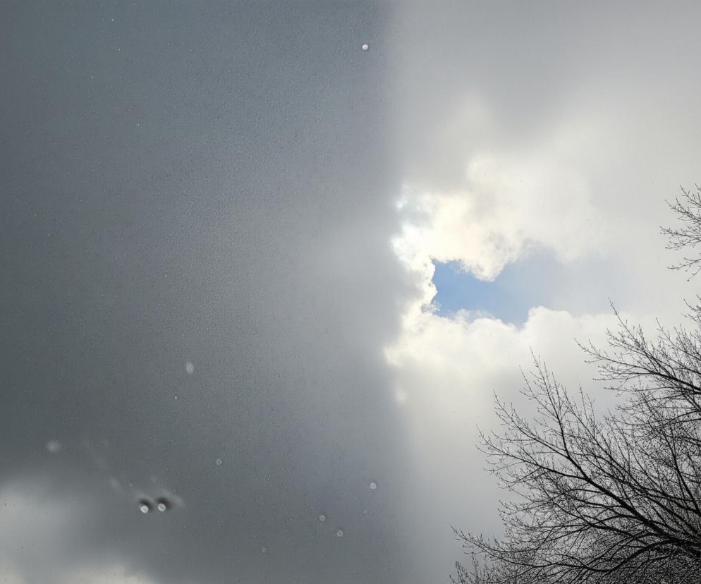 Kadr przedstawia niebo podzielone na dwie wyraźnie odmienne części. Po lewej stronie dominują ciemnoszare chmury, pokryte drobnymi, jasnymi punkcikami, przypominającymi krople deszczu lub płatki śniegu, z kilkoma większymi, rozmytymi okręgami. Po prawej stronie widać jaśniejsze, białe chmury, przez które przebija się fragment błękitnego nieba, tworząc nieregularny kształt. W dolnej części obrazu, na tle jasnych chmur, widoczne są gałęzie drzew pozbawione liści, o ciemnej, wyraźnej strukturze.