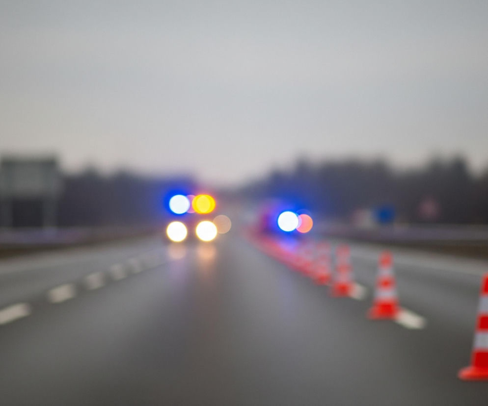 Droga asfaltowa z dwoma pasami ruchu biegnie przez środek kadru, widoczna z perspektywy jadącego samochodu. Na pasie bliżej prawej krawędzi obrazu stoi rząd czerwono-białych pachołków drogowych, które stają się coraz wyraźniejsze w miarę zbliżania się do pierwszego planu. W oddali, na przeciwległym pasie, widoczne są rozmyte światła samochodów: pulsujące niebieskie światła awaryjne oraz żółte i białe światła pojazdów. Tło stanowi zamglone, szare niebo i ciemny, rozmazany horyzont drzew lub zabudowań.