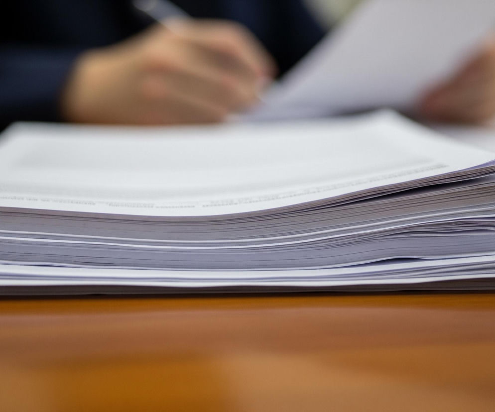 Na pierwszym planie, w dolnej części obrazu, znajduje się brązowy drewniany blat z połyskiem, na którym stoi stos białych kartek papieru. Kartki są ułożone nierówno, a niektóre krawędzie są delikatnie wygięte. W tle, częściowo rozmazany, widoczna jest osoba w ciemnej marynarce, która trzyma w prawej ręce długopis i pisze na jednej z kartek, leżącej na blacie. Obraz ma płytką głębię ostrości, z wyraźnym focusem na stosie dokumentów, podczas gdy tło jest rozmyte.