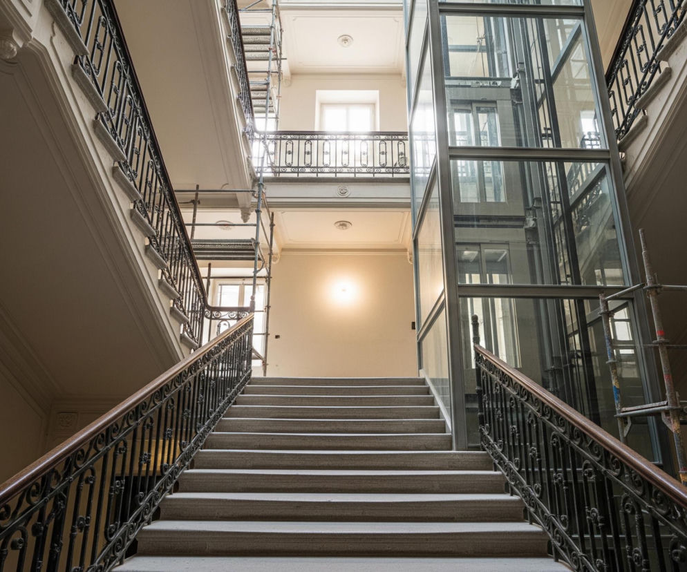 Wnętrze klatki schodowej z wysokim sufitem i oświetleniem naturalnym. Centralnie, szerokie schody prowadzą w górę, a ich stopnie są wykonane z jasnego kamienia. Po obu stronach schodów biegną balustrady z czarnego, kutego żelaza z ozdobnymi wzorami i drewnianymi poręczami w ciemnobrązowym kolorze. Na wprost, na końcu schodów, widoczna jest ściana z pojedynczym, jasnym punktem świetlnym. Po prawej stronie schodów znajduje się przeszklona kabina windy, jej metalowa konstrukcja jest widoczna przez przezroczyste panele. W tle, na wyższych kondygnacjach, widać okna, z których wpada światło, oraz elementy rusztowania budowlanego. Sufity i ściany są w jasnym, kremowym odcieniu, zdobione gzymsami.