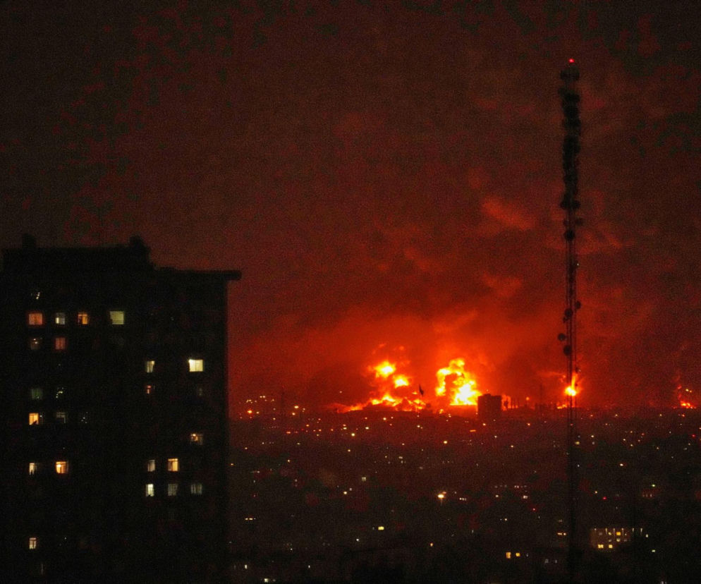 Pożary i eksplozje na Bliskim Wschodzie w nocy, z widocznymi budynkami i anteną telekomunikacyjną na pierwszym planie, ilustrujące eskalację konfliktu Izrael-Iran i wpływ na ceny ropy. Więcej o wojnie na Super Biznes.