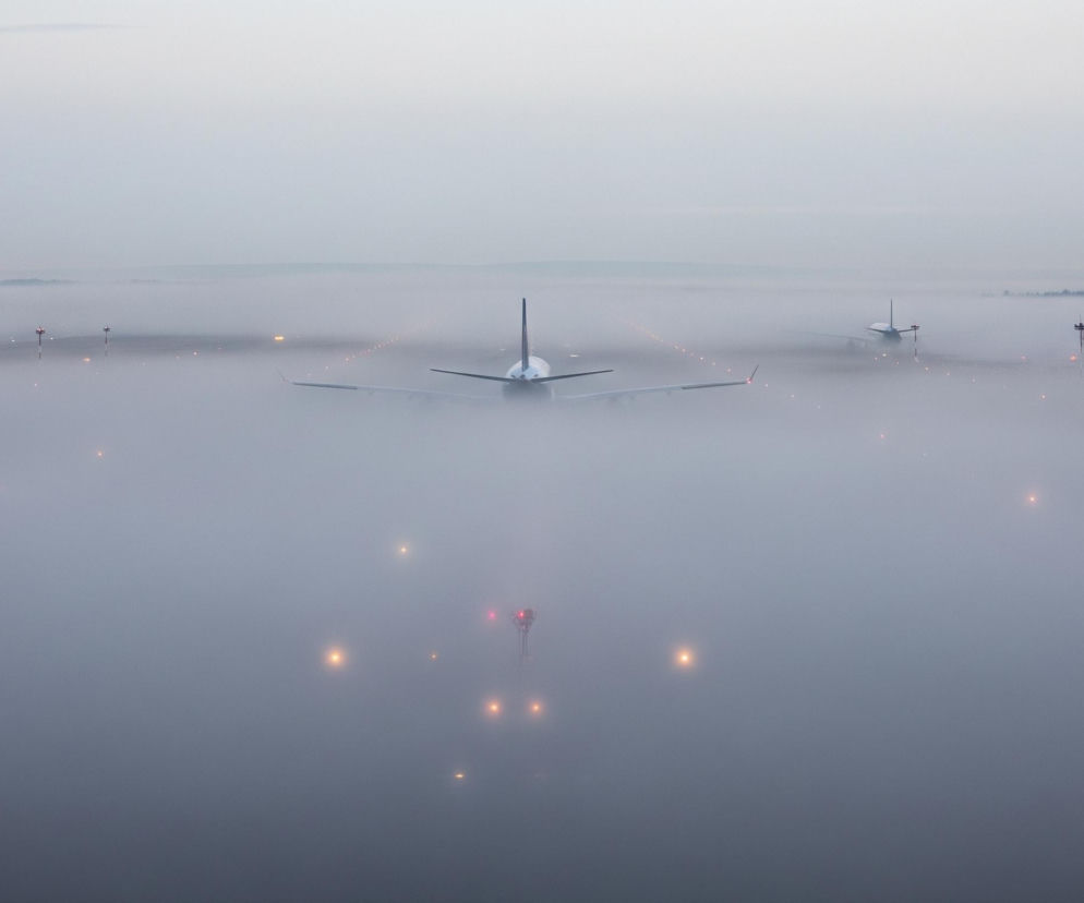 Trzy samoloty stoją na pasie startowym lotniska, otoczone gęstą mgłą o szarym zabarwieniu, która zalega również w tle, rozświetlona przez liczne pomarańczowo-czerwone światła. Największy samolot, usytuowany centralnie, zwrócony jest tyłem do widza, prezentując ciemny ogon oraz rozłożone skrzydła. Po prawej stronie w oddali, przez mgłę, widać dwie mniejsze maszyny, również zwrócone tyłem, a ich kontury są mniej wyraźne. Na horyzoncie, powyżej mgły, wyłania się delikatnie zarys ciemniejszego pasa ziemi z drzewami i kilkoma słupami, natomiast górna część nieba jest jaśniejsza i jaśniejsza, z subtelnymi szarymi chmurami.