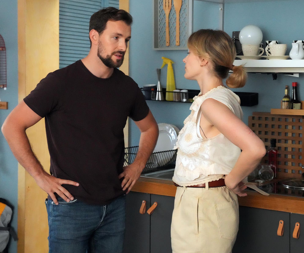 Mężczyzna z brodą i kobieta z blond włosami spiętymi w kucyk stoją w kuchni i rozmawiają. Na zdjęciu widać fragment blatu z naczyniami oraz półki z kuchennymi akcesoriami. Renata (Anna Mrozowska) i Marcin (Oskar Stoczyński) na tle kuchni w serialu Barwy szczęścia. Więcej o ich losach na stronie Super Seriale.