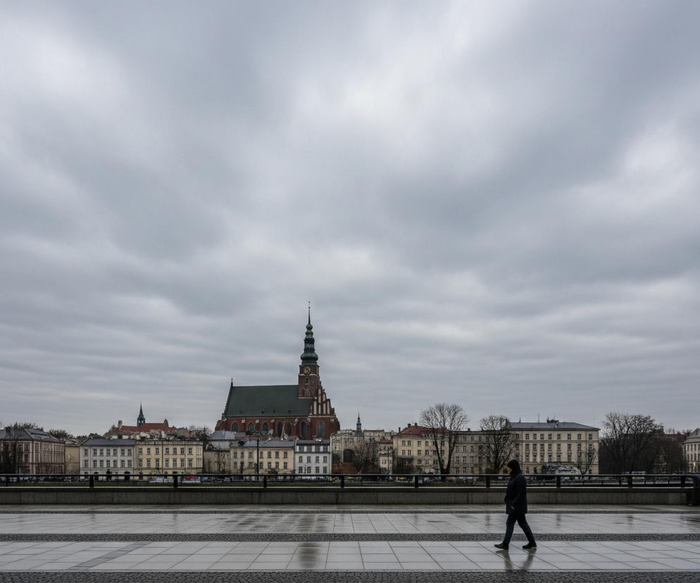 Samotna postać idzie prawą stroną dużego, jasnoszarego chodnika z widocznymi mokrymi plamami odbijającymi światło. W tle, za ciemnym, metalowym ogrodzeniem, rozciąga się miejski krajobraz z dominującym, wysokim kościołem z czerwoną cegłą i zielonym dachem, zwieńczonym spiczastą wieżą zegarową. Dookoła kościoła stoją liczne, klasyczne budynki o jasnych elewacjach i ciemnych dachach, a za nimi widać nagie drzewa, sugerujące chłodniejszą porę roku. Całość panoramy przykrywa jednolicie zachmurzone, szare niebo.