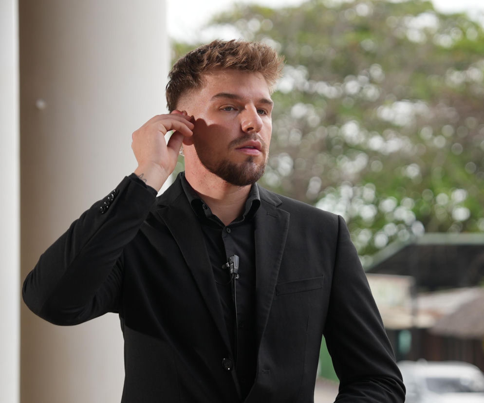 Uczestnik Darek z programu Hotel Paradise w czarnym garniturze z mikroportem, z ręką dotykającą ucha. Tło to rozmyta zieleń i fragmenty budynku. Zdjęcie ilustruje artykuł o kontrowersyjnym zachowaniu Darka w programie Hotel Paradise, dostępne na stronie Super Seriale.