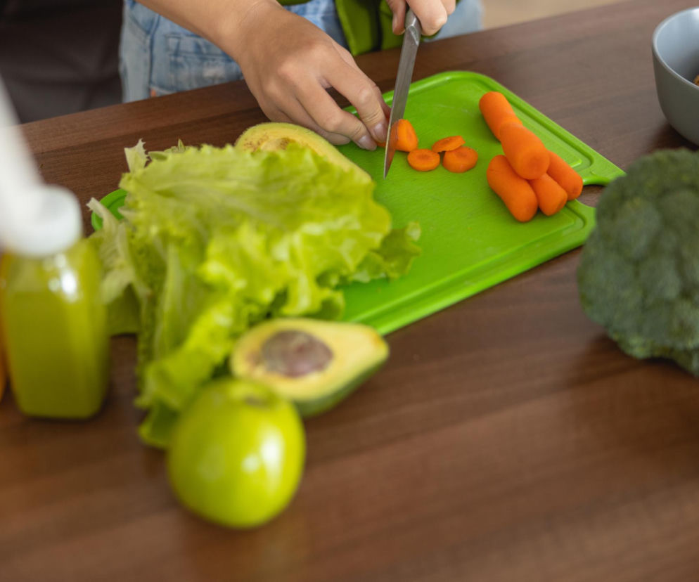 Osoba krojąca marchewkę na zielonej desce, obok awokado, sałata, jabłko i napoje. Zdjęcie ilustruje problem ślizgającej się deski, który może prowadzić do skaleczeń i bólu karku. Dowiedz się więcej o tym, jak zabezpieczyć deskę i chronić swoje zdrowie na Poradnik Zdrowie.