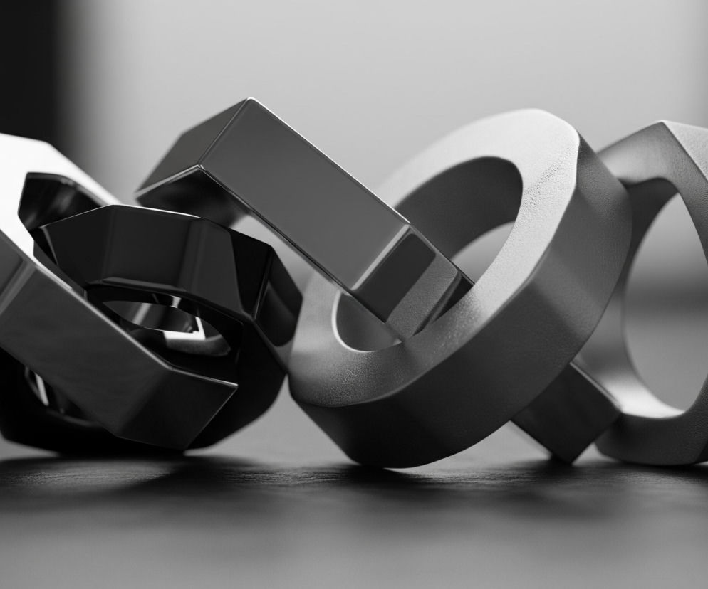 Na ciemnym, matowym tle widoczne są trzy połączone ze sobą abstrakcyjne, geometryczne kształty. Obiekty te, przypominające metalowe ogniwa, mają odcienie szarości i czerni, z błyszczącymi refleksami światła na niektórych powierzchniach. Dwa z nich, te po lewej, są gładkie i odblaskowe, a ich krawędzie są ostre i precyzyjne. Trzeci obiekt po prawej ma matową, chropowatą teksturę. Całość spoczywa na ciemnej, delikatnie teksturowanej powierzchni, rzucając na nią subtelne cienie.