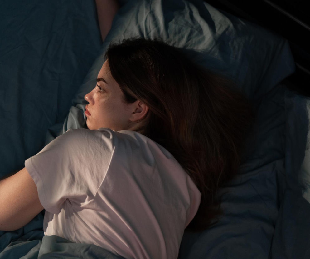 Kobieta z otwartymi oczami leży w łóżku na granatowej pościeli, jej twarz oświetlona ciepłym światłem. Wygląda na zmartwioną lub zestresowaną, co odzwierciedla problem z zasypianiem. Na portalu Poradnik Zdrowie znajdziesz sposoby na obniżenie kortyzolu i poprawę snu.