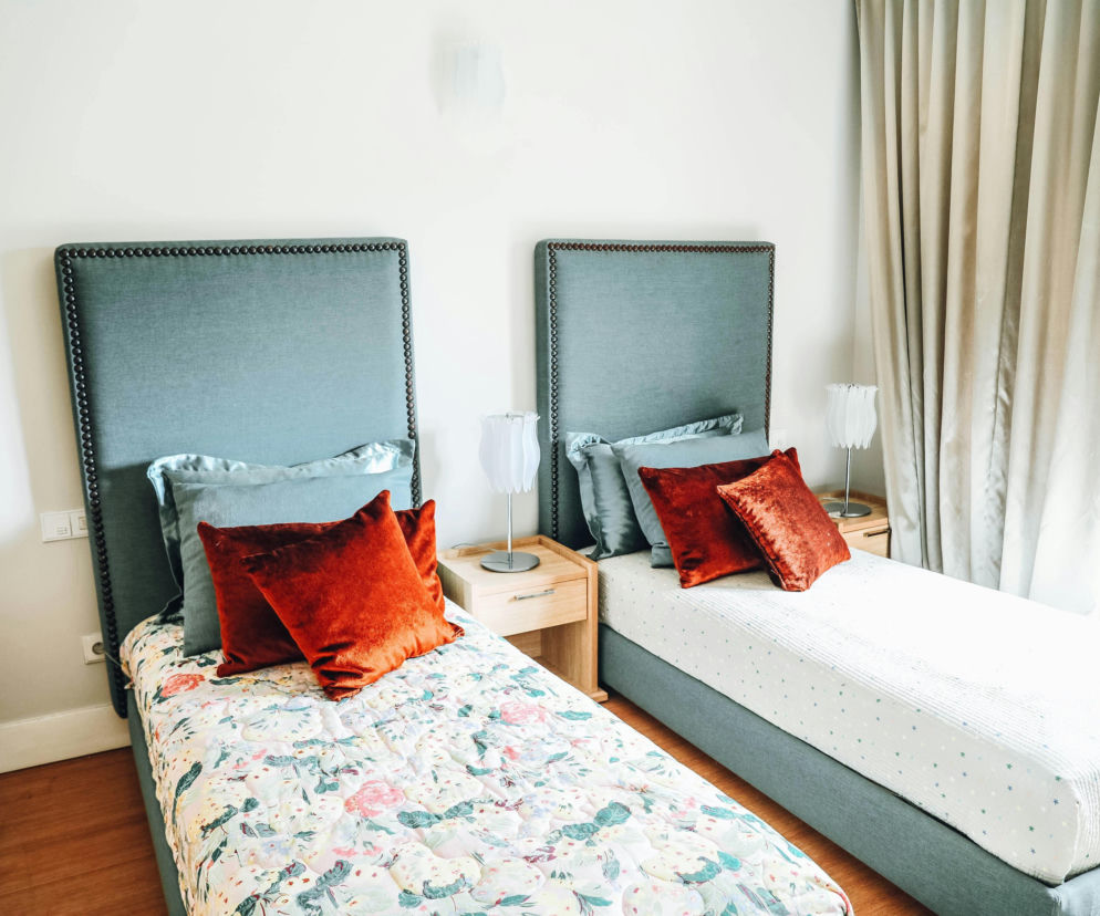 Dwa oddzielne łóżka z szarymi zagłówkami i różnymi pościelami w jasnej sypialni, obok których stoją szafki nocne z lampkami. To rozwiązanie staje się coraz popularniejsze dla par, które chcą dbać o zdrowie, o czym przeczytasz na Poradnik Zdrowie.