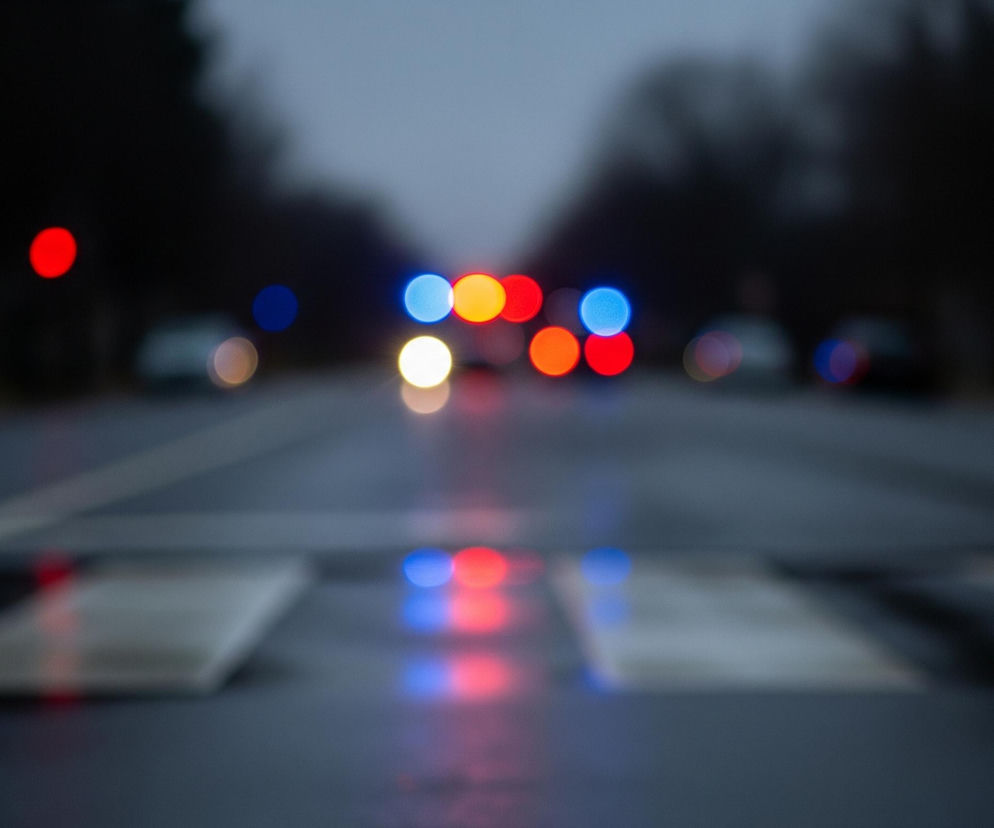 Rozmyty widok ulicy nocą lub wczesnym rankiem. Na pierwszym planie widać ciemny, mokry asfalt z zamazanymi białymi pasami przejścia dla pieszych. W oddali, wzdłuż osi drogi, rozciągają się drzewa po obu stronach. Głównym elementem są liczne, kolorowe, rozmyte światła samochodów: dominują jaskrawe czerwienie, błękity, pomarańcze i biele, odbijające się częściowo na mokrej nawierzchni ulicy.