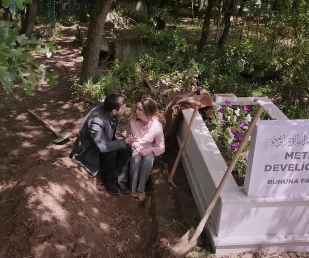 Cihan i Hancer na cmentarzu, siedzą w wykopanym grobie obok nagrobka z napisem Metin Develioğlu. Obok nich leżą dwie łopaty. W tle widać drzewa i zarośla. Scena z serialu Panna młoda, o której więcej dowiesz się na Super Seriale.