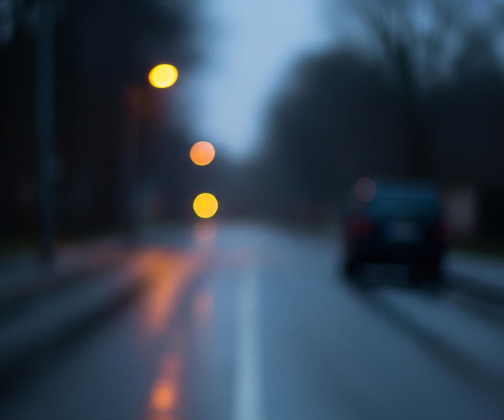 Droga pokryta deszczem rozciąga się w głąb obrazu, z wyraźną białą linią przerywaną pośrodku, a jej mokra powierzchnia odbija rozmyte, pomarańczowe światła. Po lewej stronie widać słup, a na tle ciemnego, rozmytego krajobrazu wznoszą się trzy okrągłe, jaskrawożółte punkty światła, prawdopodobnie latarnie uliczne. Po prawej stronie, w oddali, stoi rozmazany, ciemny samochód z widocznymi czerwonymi światłami. Cała scena jest zamglona i utrzymana w ciemnych, niebieskawo-szarych barwach.