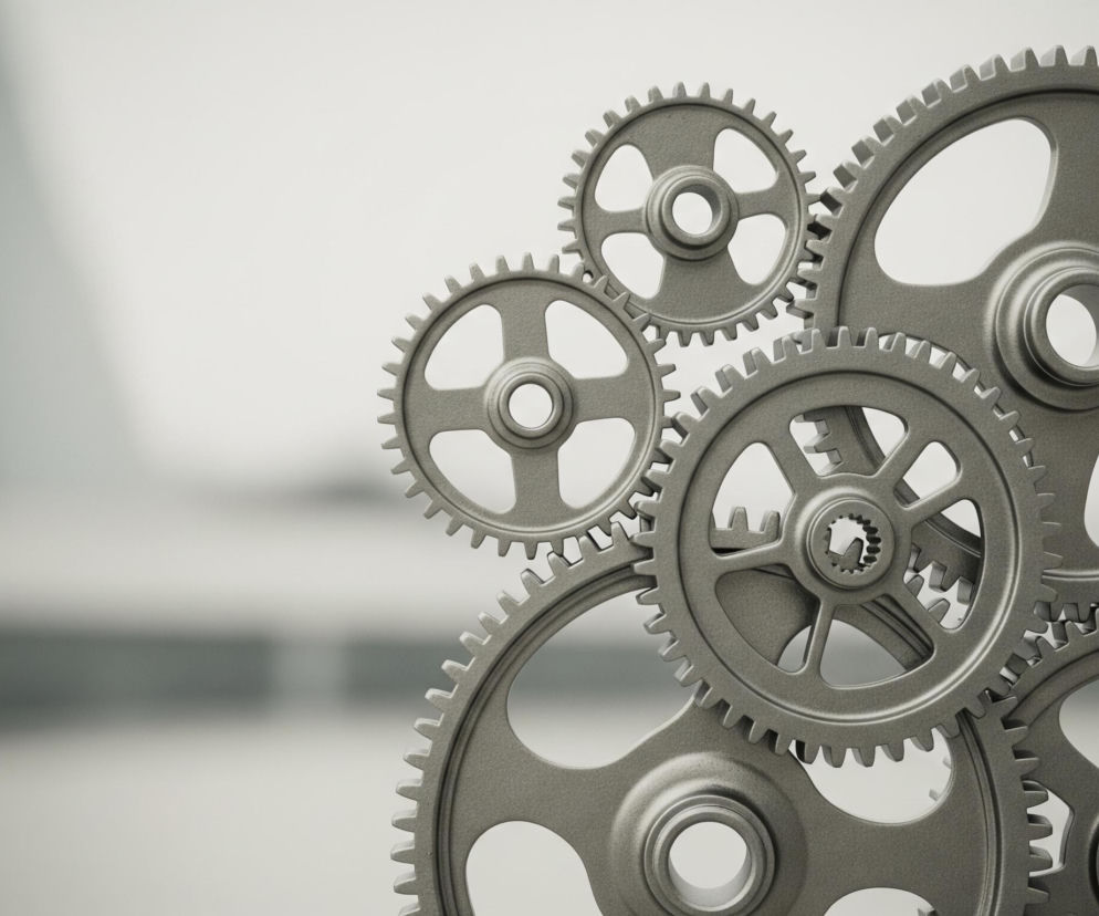 Na zdjęciu dominują szare metalowe koła zębate, ułożone w zwartej grupie po prawej stronie. Widocznych jest siedem kół o różnej wielkości, z których największe znajdują się na dole i po prawej, a mniejsze na górze i w środku kompozycji. Wszystkie koła zębate są ze sobą połączone, ich zęby zazębiają się nawzajem. Tło jest rozmyte, w odcieniach szarości i bieli, tworząc efekt przestrzeni za kołami zębatymi.