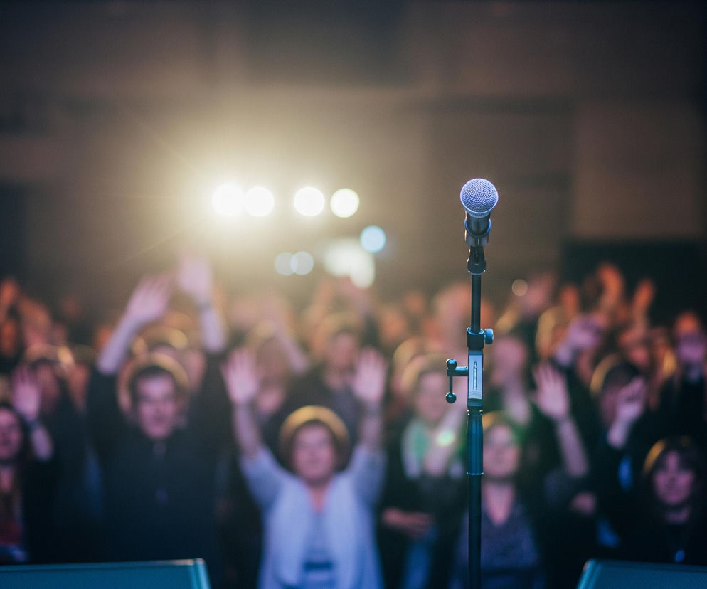 Na pierwszym planie, centralnie, stoi czarny mikrofon na statywie, skierowany do przodu. Mikrofon ma srebrzystą, siateczkową główkę i ciemny korpus, lekko oświetlony od przodu. W tle, rozmyty tłum ludzi, wielu z nich ma podniesione ręce w geście aplauzu lub interakcji, z niewyraźnymi rysami twarzy. Za tłumem widać jasne, rozproszone źródła światła, tworzące efekt bokeh. Całość jest utrzymana w ciemnej tonacji z ciepłym, żółtawo-pomarańczowym oświetleniem w tle i chłodniejszym, niebieskawym akcentem na mikrofonie.