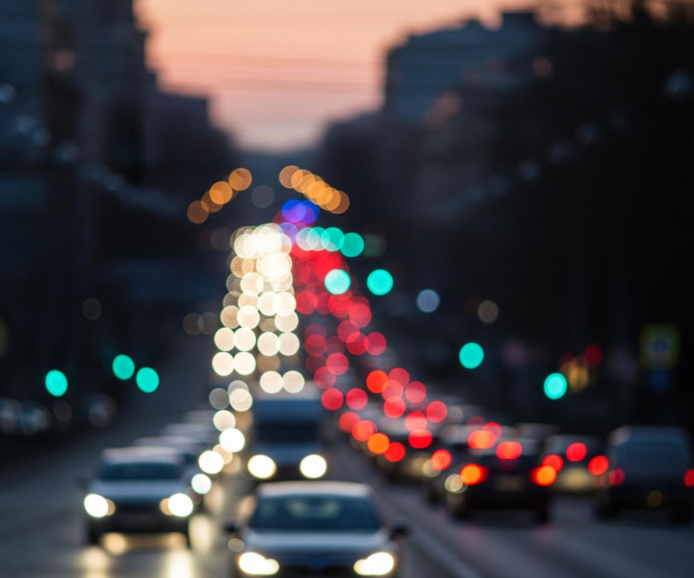 Rozmazany obraz przedstawia ruch uliczny o zmierzchu lub w nocy, z licznymi światłami samochodów tworzącymi efekt bokeh. Na pierwszym planie widoczne są niewyraźne sylwetki samochodów, z których niektóre mają włączone jasne, białe światła z przodu, a inne czerwone światła z tyłu. Ulicę po lewej stronie oświetlają rozmyte, turkusowe światła sygnalizacji. Horyzont na górze obrazu rozświetla pomarańczowo-różowa poświata zachodzącego słońca, kontrastując z ciemnymi sylwetkami budynków.