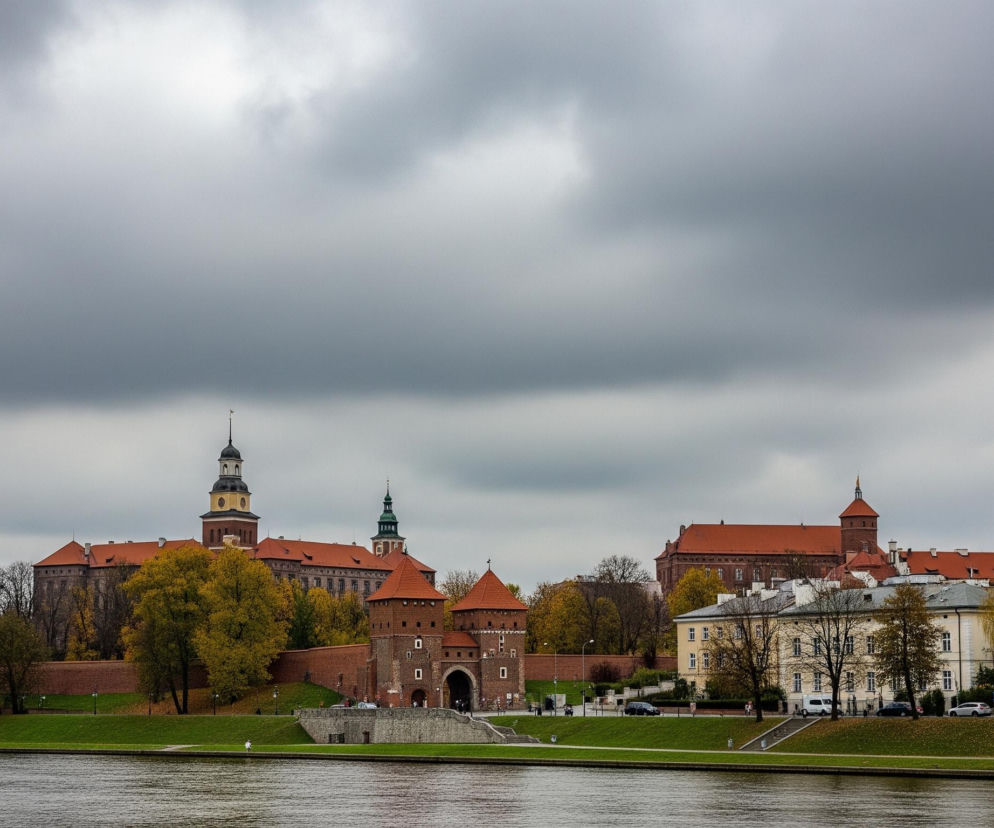 Szeroki kadr przedstawia rzekę na pierwszym planie, z brzegiem pokrytym zieloną trawą po prawej i lewej stronie. Wzdłuż prawego brzegu rzeki ciągnie się rząd kamienic o jasnych elewacjach i pomarańczowych dachach, a przed nimi zaparkowane są samochody. Na lewym brzegu wznosi się kompleks budowli z cegły, z wysokimi murami obronnymi, czterema wieżami o stożkowych dachach oraz dwoma większymi wieżami zakończonymi kopułami, z których jedna ma żółte elementy. Całość otaczają drzewa, niektóre z jasnożółtymi liśćmi, inne pozbawione liści, a nad miastem rozciąga się zachmurzone, szare niebo.
