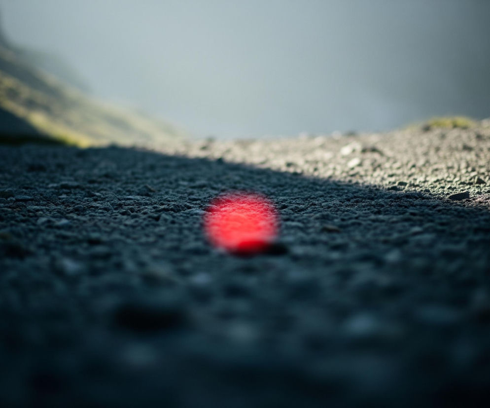 Centralnie w kadrze, na pierwszym planie, znajduje się rozmyta, jasnoczerwona plama światła. Otacza ją ciemny, żwirowy teren, częściowo oświetlony blaskiem z prawej strony, tworząc wyraźną linię cienia. W oddali, rozmyte i niewyraźne, widoczne są zarysy wzniesień pokrytych zieloną roślinnością, a powyżej nich mgliste, jasnoszare niebo.