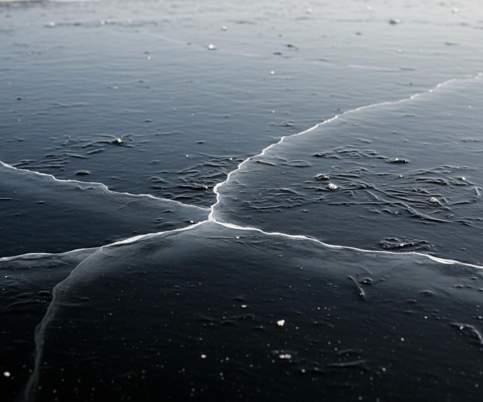 Ciemną, lodową powierzchnię poprzecinaną wyraźnymi pęknięciami. Lód jest w większości gładki i ma barwę od ciemnogranatowej do czarnej w dolnej części, przechodzącą w jaśniejszy, mleczny odcień w górnej części kadru. Białe linie pęknięć rozchodzą się promieniście od centralnego punktu w dolnej części obrazu, tworząc trójkątny wzór. Na powierzchni lodu widoczne są drobne, nieregularne białe plamki i bąbelki, a także subtelne, faliste tekstury.