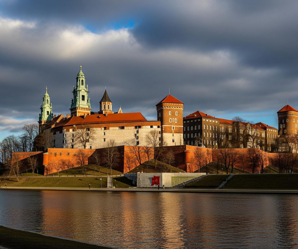 Na pierwszym planie widoczna jest osoba w ciemnej, pikowanej kurtce z futrzanym kołnierzem i czarnej czapce, idąca brukowaną ścieżką wzdłuż rzeki. Rzeka zajmuje większą część dolnej połowy kadru, a jej powierzchnia odbija pomarańczowe światło zachodzącego słońca oraz fragmenty nieba i zabudowań. Po drugiej stronie rzeki dominuje kompleks średniowiecznych budowli z ceglanymi murami obronnymi i basztami o czerwonobrunatnych dachach, otoczony bezlistnymi drzewami i zielonymi wzgórzami. Nad budynkami rozpościera się częściowo zachmurzone niebo z widocznymi błękitnymi fragmentami.