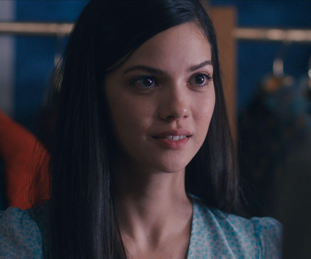 Portret młodej kobiety o długich, ciemnych włosach, delikatnie uśmiechającej się i patrzącej w bok, w stylizowanej sukience w drobne, jasne wzory. To fotografia Eylül Kandemir, aktorki grającej Füsun w serialu Muzeum niewinności, o którym przeczytasz więcej na Super Seriale.