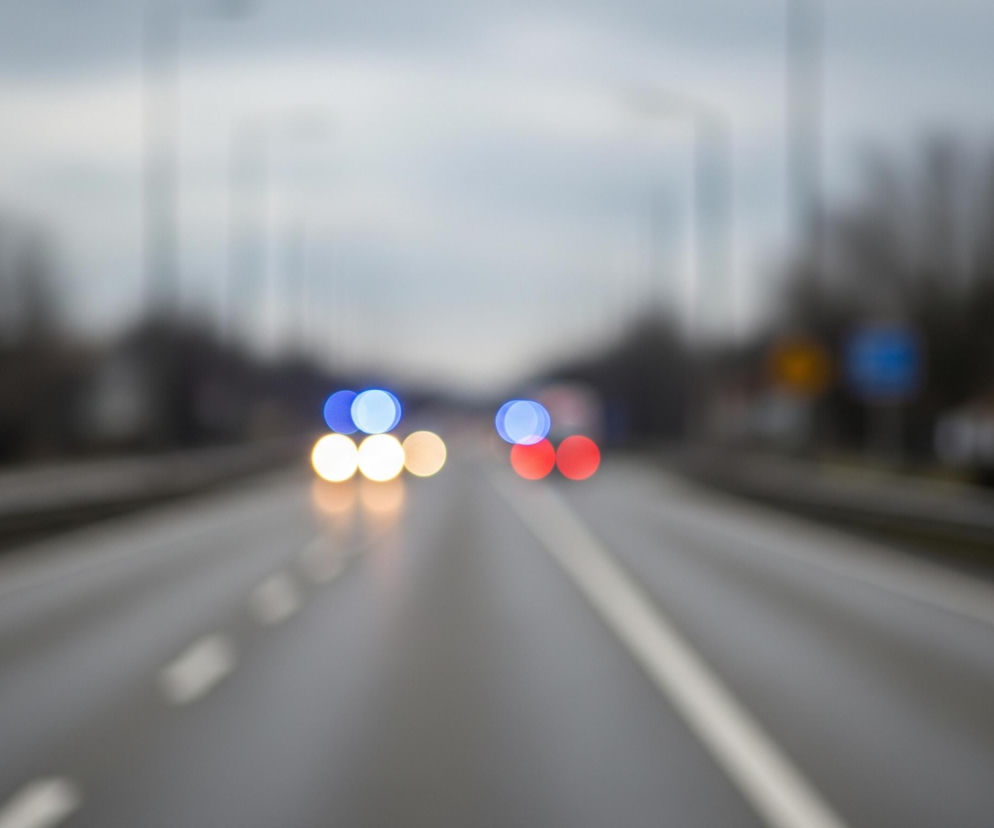 Droga z szarym asfaltem, oświetlona rozmytymi światłami samochodów, biegnie przez centrum kadru. Na jej powierzchni widoczne są białe linie, tworzące pasy ruchu oraz strzałki. W tle widać niewyraźne, rozmyte kształty budynków oraz drzew pod szarym, zachmurzonym niebem. Na środku drogi znajdują się rozmyte światła pojazdów: białe i żółte z przodu, a za nimi po prawej stronie dwa czerwone światła, po lewej zaś jedno niebieskie.