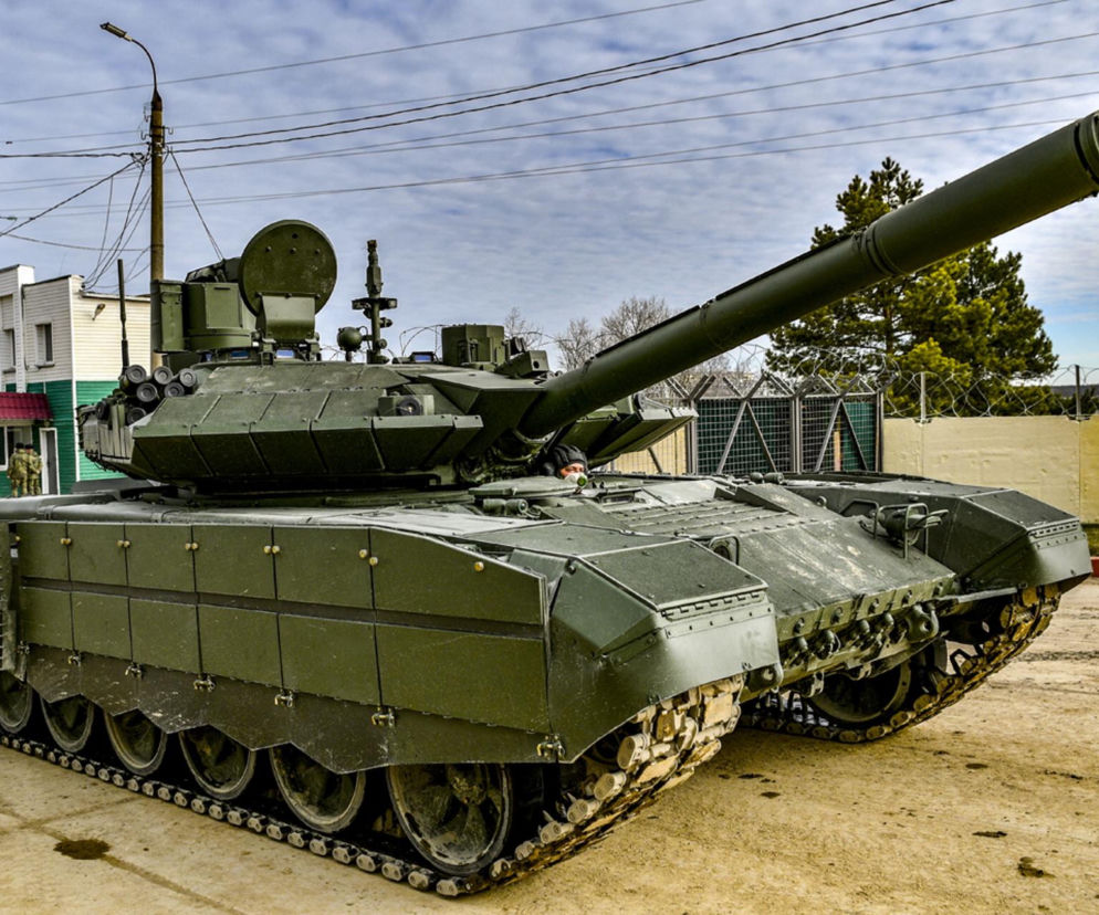 Zielony czołg T-90M Proryw z długą lufą i charakterystycznym kamuflażem, widoczny pod pochmurnym niebem, obok budynku i słupa wysokiego napięcia. To symbol modernizacji rosyjskiej armii, o której przeczytasz więcej na naszym portalu.